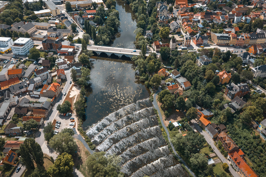 Aerial view of the Saale river flowing through the town of Saalfeld featuring a stepped weir a stone bridge historic buildings and lush green trees in Saalfeld Thuringia Germany. Aerial view of the Saale river flowing through the town of Saalfeld featuring a stepped weir a stone bridge historic buildings and lush green trees in Saalfeld Thuringia Germany.