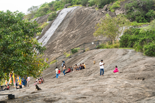 Sri Lanka 21 February 2026 View of people sitting on a large sloping rock face near a thin waterfall surrounded by lush green trees and Buddhist flags.