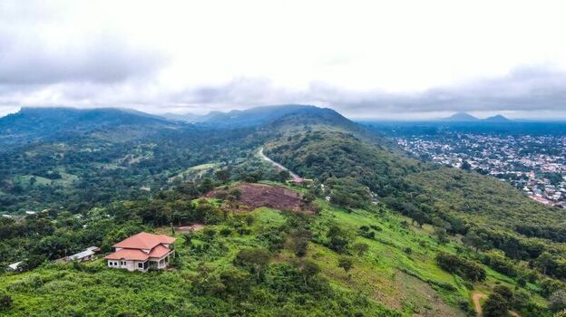 Aerial view of a house nestled on a lush green hillside with a winding road leading towards a distant town under a cloudy sky in Ghana. Aerial view of a house nestled on a lush green hillside with a winding road leading towards a distant town under a cloudy sky in Ghana.