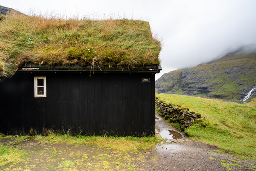 View of a traditional black wooden house with a grass roof situated in a lush green landscape with a stone wall misty mountains and a waterfall in the background Faroe Islands.