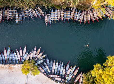 Aerial view of vibrant boats line the tranquil riverbanks near lush greenery contrasting with the dark water and a lone swimmer Kineret Israel.