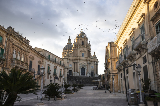 View of the Duomo di San Giorgio with birds flying over the baroque facade and historic stone buildings in Ragusa Sicily Italy.