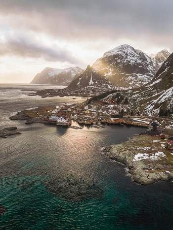 Aerial view of sunlight piercing through the clouds illuminating the snow dusted mountains and the tranquil waters surrounding the village Tind Nordland Norway.