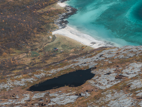 Aerial view of the rugged coastline of Senja featuring a small red cabin near a white sand beach turquoise waters and rocky terrain in Senja Troms Norway.