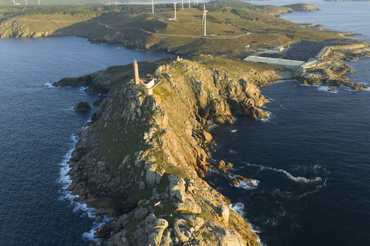 Aerial view of a lighthouse perched on a rocky peninsula surrounded by the Atlantic Ocean with wind turbines on the grassy hills in the background in Galicia Spain.