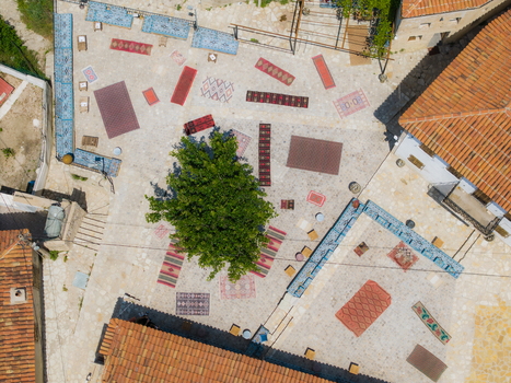 Aerial view of a traditional stone courtyard decorated with colorful patterned rugs and a central green tree surrounded by terracotta tiled roofs in Dhermi Vlore County Albania. Aerial view of a traditional stone courtyard decorated with colorful patterned rugs and a central green tree surrounded by terracotta tiled roofs in Dhermi Vlore County Albania.