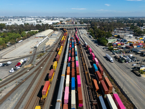 Aerial view of a busy railyard filled with colorful shipping containers on freight trains with industrial warehouses and nearby roads in Carson California United States.
