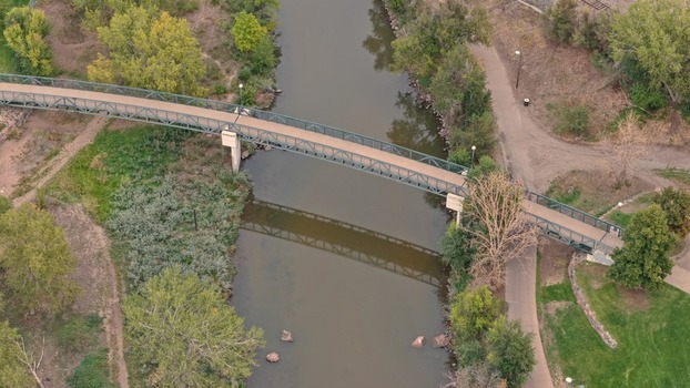 Aerial view of a pedestrian bridge spanning a river in a park setting with surrounding trees and pathways in Denver Colorado United States. Aerial view of a pedestrian bridge spanning a river in a park setting with surrounding trees and pathways in Denver Colorado United States.