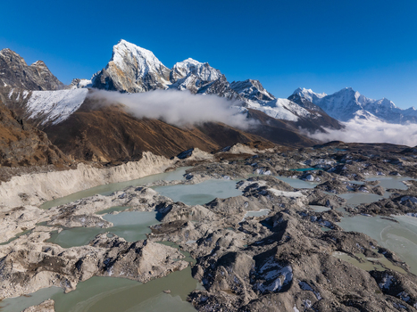 Aerial view of the rugged Himalayan glacial landscape featuring snow capped mountain peaks rocky moraine and turquoise meltwater pools under a clear blue sky in Nepal.