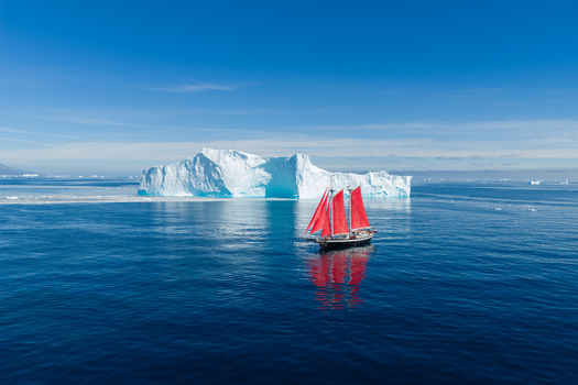 Aerial view of a sailboat with striking red sails navigating through deep blue Arctic waters past a massive iceberg under a clear sky in Greenland. Aerial view of a sailboat with striking red sails navigating through deep blue Arctic waters past a massive iceberg under a clear sky in Greenland.