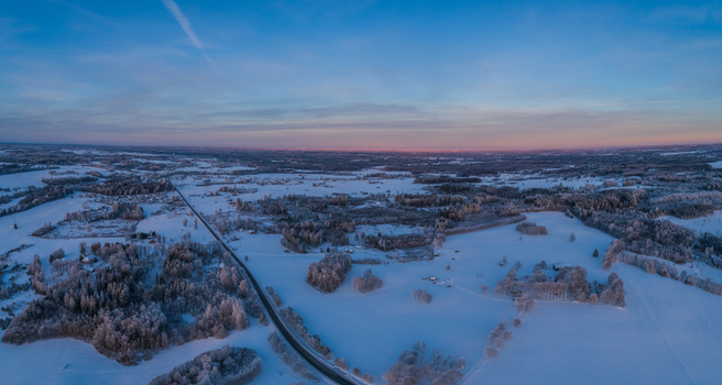 Aerial view of a vast snow covered landscape featuring a winding road cutting through frost covered forests and open fields under a soft twilight sky in Latvia. Aerial view of a vast snow covered landscape featuring a winding road cutting through frost covered forests and open fields under a soft twilight sky in Latvia.