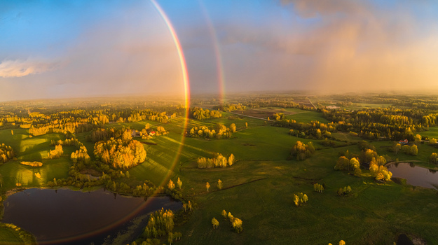 Aerial view of a double rainbow arching over a lush green rural landscape with scattered trees and a small pond illuminated by golden hour light in Latvia. Aerial view of a double rainbow arching over a lush green rural landscape with scattered trees and a small pond illuminated by golden hour light in Latvia.