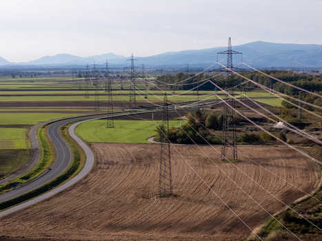 Aerial view of high voltage power lines and electricity pylons stretching across green fields and a winding road with mountains in the distance Slovenia. Aerial view of high voltage power lines and electricity pylons stretching across green fields and a winding road with mountains in the distance Slovenia.