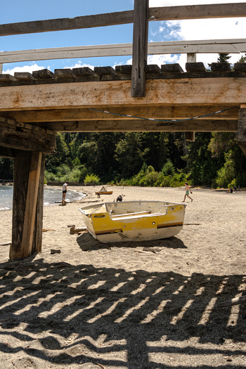 View of Bahia Brava with a yellow boat on the sandy beach under a wooden pier with people walking along the shore in Villa La Angostura Neuquen Province Argentina. View of Bahia Brava with a yellow boat on the sandy beach under a wooden pier with people walking along the shore in Villa La Angostura Neuquen Province Argentina.