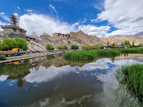 View of Shey Palace Ladakh reflecting in a calm pond surrounded by green trees and rocky mountains under a blue sky with clouds in Shey Ladakh India.