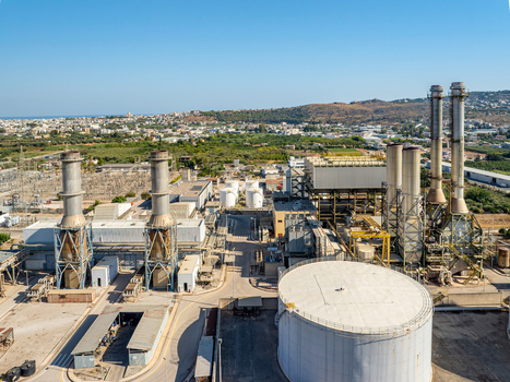 Aerial view of the industrial power plant facility featuring large storage tanks tall smokestacks and electrical infrastructure set against the landscape of Chania Greece.
