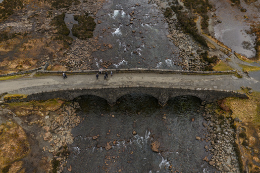 Aerial view of the historic Sligachan Old Bridge spanning a rocky river with people walking across the stone structure in the rugged Highlands Sligachan Scotland United Kingdom. Aerial view of the historic Sligachan Old Bridge spanning a rocky river with people walking across the stone structure in the rugged Highlands Sligachan Scotland United Kingdom.