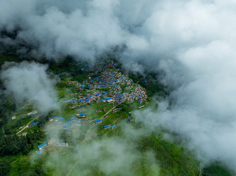 Aerial view of a mountain village with blue roofed houses nestled among lush green terraced fields and surrounded by thick clouds in Nepal.