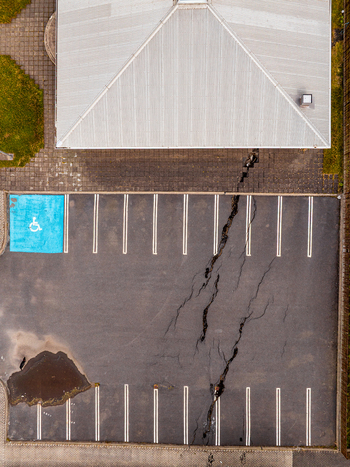 Aerial view of a large fissure splitting the asphalt surface of a parking lot with a blue handicap parking space and a puddle in Iceland. Aerial view of a large fissure splitting the asphalt surface of a parking lot with a blue handicap parking space and a puddle in Iceland.