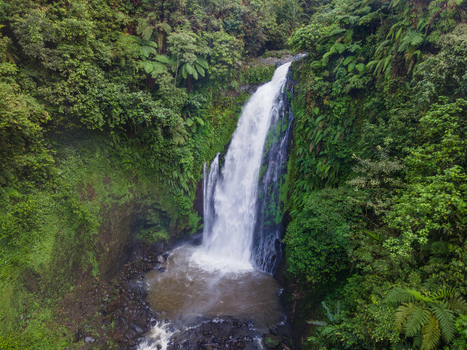 Aerial view of Gomblang Waterfall cascading down a rocky cliff surrounded by lush tropical forest and dense green foliage in Kecamatan Kedungbanteng Jawa Tengah Indonesia. Aerial view of Gomblang Waterfall cascading down a rocky cliff surrounded by lush tropical forest and dense green foliage in Kecamatan Kedungbanteng Jawa Tengah Indonesia.