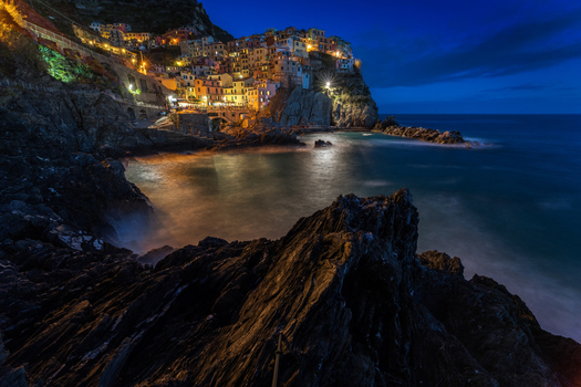View of the illuminated colorful houses of Manarola perched on a cliff overlooking the sea at twilight with dark rocks in the foreground Manarola Liguria Italy. View of the illuminated colorful houses of Manarola perched on a cliff overlooking the sea at twilight with dark rocks in the foreground Manarola Liguria Italy.