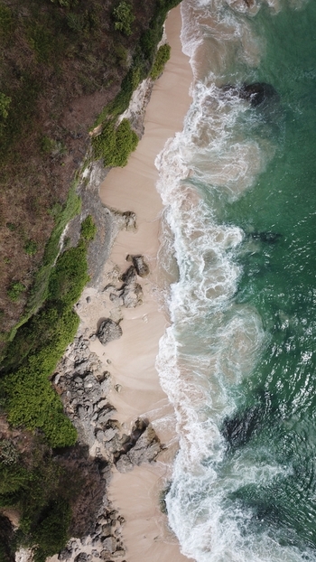 Aerial view of a sandy beach with rocky cliffs and lush greenery meeting the turquoise ocean waves in Indonesia.