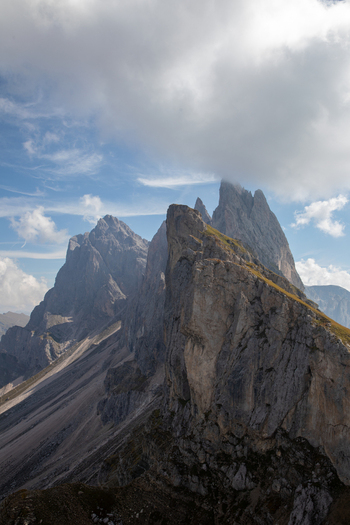 View of the jagged peaks of the Dolomites with steep rock faces and grassy ridges under a cloudy blue sky in Rocca Pietore Veneto Italy. View of the jagged peaks of the Dolomites with steep rock faces and grassy ridges under a cloudy blue sky in Rocca Pietore Veneto Italy.