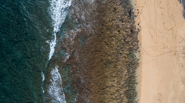 Aerial view of the coastline featuring turquoise waves crashing against a rocky reef and a sandy beach with visible footprints in Bajawa Nusa Tenggara Timur Indonesia. Aerial view of the coastline featuring turquoise waves crashing against a rocky reef and a sandy beach with visible footprints in Bajawa Nusa Tenggara Timur Indonesia.