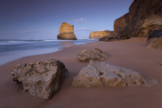 View of the limestone stacks of the Twelve Apostles Marine National Park with large foreground rocks on a sandy beach at twilight in Australia. View of the limestone stacks of the Twelve Apostles Marine National Park with large foreground rocks on a sandy beach at twilight in Australia.