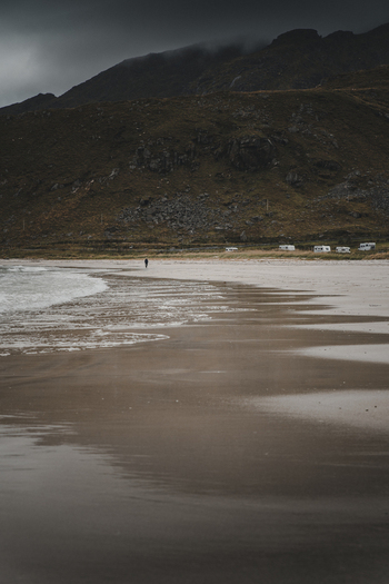 View of Haukland Beach with a person walking along the wet sand dramatic mountains and distant campers under an overcast sky in Leknes Nordland Norway. View of Haukland Beach with a person walking along the wet sand dramatic mountains and distant campers under an overcast sky in Leknes Nordland Norway.