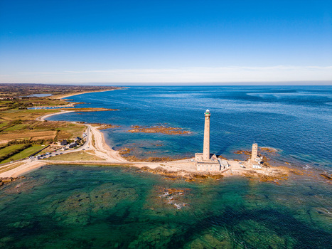 Aerial view of Gatteville Lighthouse standing on a rocky point along the coastline with clear blue sea and green fields in the background in Gatteville le Phare Normandie France.