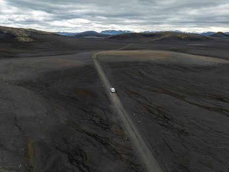 Aerial view of a white car driving on a gravel road through a vast dark volcanic landscape under a cloudy sky in Iceland. Aerial view of a white car driving on a gravel road through a vast dark volcanic landscape under a cloudy sky in Iceland.
