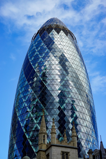 View of 30 St Mary Axe also known as The Gherkin with its distinctive glass facade rising above historic stone architecture against a blue sky in London England United Kingdom.