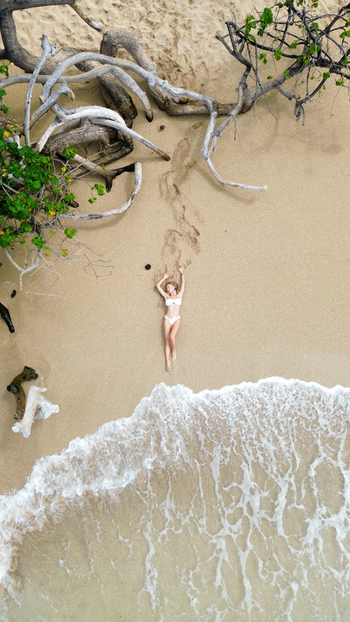 Aerial view of a woman in a white bikini lying on the sand at Waikiki Beach surrounded by driftwood and gentle ocean waves in Honolulu Hawaii United States.
