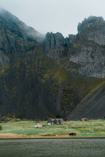View of Stokksnes with the dramatic jagged peaks of Vestrahorn mountain rising above small houses with red roofs in a grassy coastal landscape in Iceland.