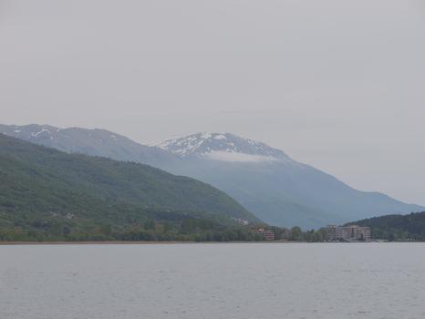 View of the calm waters of Lake Ohrid with snow capped mountains rising in the background under an overcast sky in Ohrid Municipality of Ohrid North Macedonia. View of the calm waters of Lake Ohrid with snow capped mountains rising in the background under an overcast sky in Ohrid Municipality of Ohrid North Macedonia.