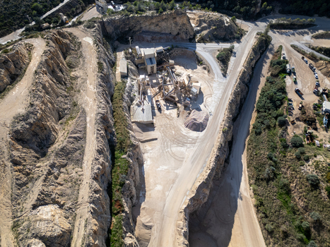 Aerial view of an open pit quarry with industrial machinery gravel piles and dirt roads surrounded by rocky cliffs and vegetation in Teulada Valencian Community Spain.
