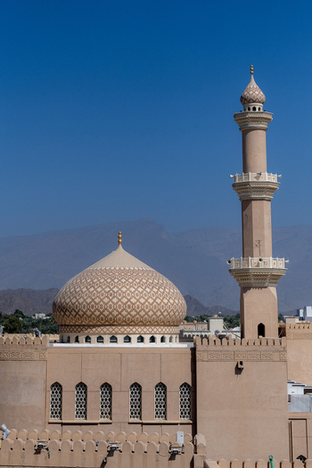 View of a mosque with an intricate patterned dome and a tall minaret set against a clear blue sky and distant mountains in Muscat Muscat Governorate Oman. View of a mosque with an intricate patterned dome and a tall minaret set against a clear blue sky and distant mountains in Muscat Muscat Governorate Oman.