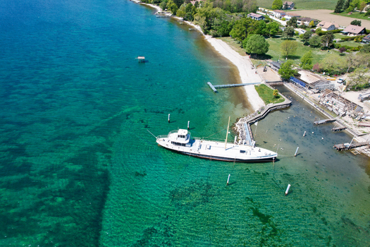 Aerial view of a white boat moored in the turquoise waters of Lake Geneva near the green shoreline and houses in Perroy Vaud Switzerland. Aerial view of a white boat moored in the turquoise waters of Lake Geneva near the green shoreline and houses in Perroy Vaud Switzerland.
