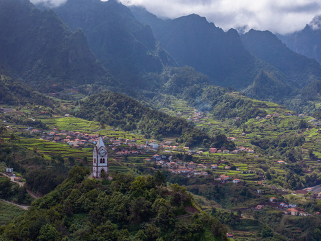 Aerial view of a historic clock tower standing on a lush green hill surrounded by terraced agricultural fields and small houses nestled in the mountainous landscape of Madeira. Aerial view of a historic clock tower standing on a lush green hill surrounded by terraced agricultural fields and small houses nestled in the mountainous landscape of Madeira.