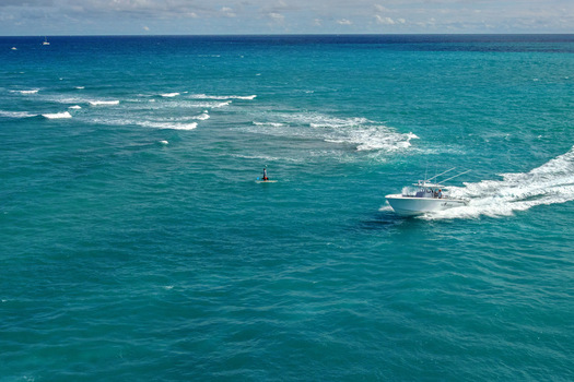 Aerial view of a boat cutting through the turquoise waters contrasting with the white wake near a solitary figure standing on a paddleboard Jupiter Florida United States.