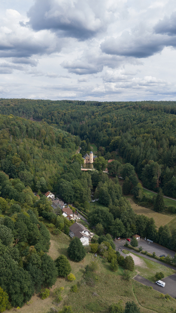 Aerial view of Schloss Mespelbrunn nestled in a lush green forest valley with surrounding trees rolling hills and a cloudy sky in Mespelbrunn Bayern Germany.