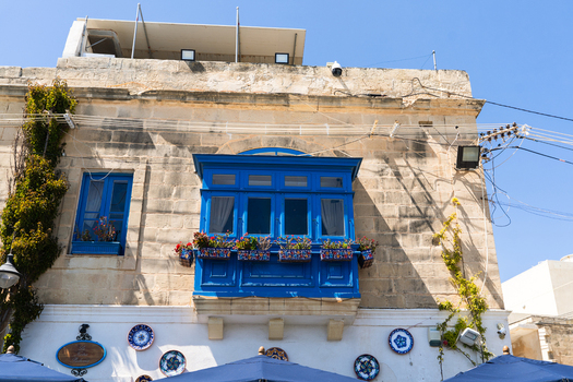View of a traditional limestone building featuring a vibrant blue wooden balcony with flower boxes and decorative ceramic plates in Marsaskala Southern Region Malta. View of a traditional limestone building featuring a vibrant blue wooden balcony with flower boxes and decorative ceramic plates in Marsaskala Southern Region Malta.