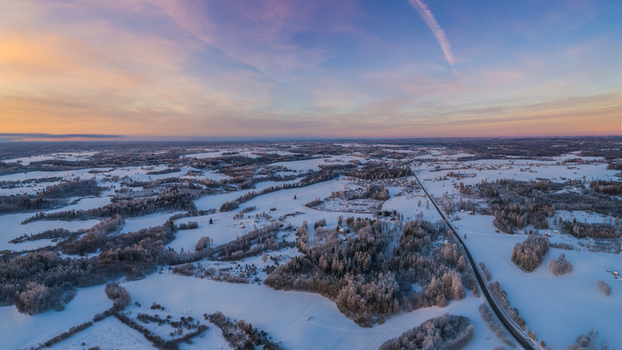Aerial view of a snow covered landscape featuring frozen fields dense forests and a winding road under a soft sunset sky in Latvia. Aerial view of a snow covered landscape featuring frozen fields dense forests and a winding road under a soft sunset sky in Latvia.