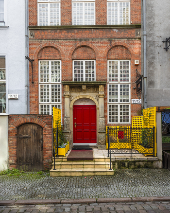 View of a historic brick building with an ornate stone doorway and a vibrant red door featuring white window frames and yellow lattice fencing in Gdansk Pomeranian Voivodeship Poland. View of a historic brick building with an ornate stone doorway and a vibrant red door featuring white window frames and yellow lattice fencing in Gdansk Pomeranian Voivodeship Poland.