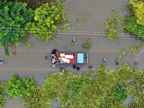 Banshkhali Upazila Bangladesh 10 July 2026 Aerial view of a truck driving through a flooded road with people wading in the water surrounded by lush green vegetation. Banshkhali Upazila Bangladesh 10 July 2026 Aerial view of a truck driving through a flooded road with people wading in the water surrounded by lush green vegetation.