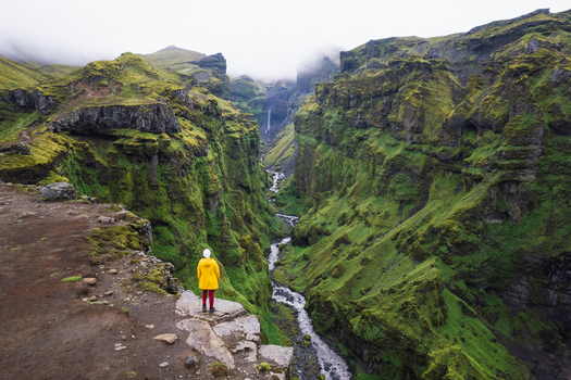Aerial view of a person in a yellow jacket standing on a cliff edge overlooking Mulagljufur Canyon with its lush green walls and a winding river Eastern Region Iceland. Aerial view of a person in a yellow jacket standing on a cliff edge overlooking Mulagljufur Canyon with its lush green walls and a winding river Eastern Region Iceland.