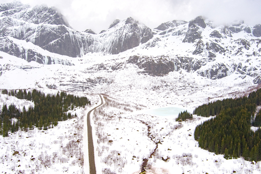 Aerial view of the snow covered Lofoten mountains with a winding road cutting through the landscape surrounded by pine forests and a frozen lake in Nordland Norway.