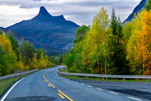 View of Stor Graddis mountain peak rising above a winding asphalt road lined with vibrant autumn trees in Saltdal Nordland Norway.