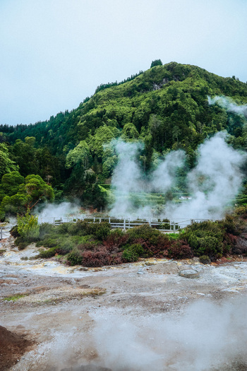 View of ethereal steam rising from the geothermal landscape contrasting with the verdant hills and dark foliage creating a mystical atmosphere Furnas Azores Portugal.